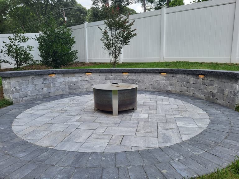 Modern gas fire pit on a paver patio with a lit seat wall at dusk
