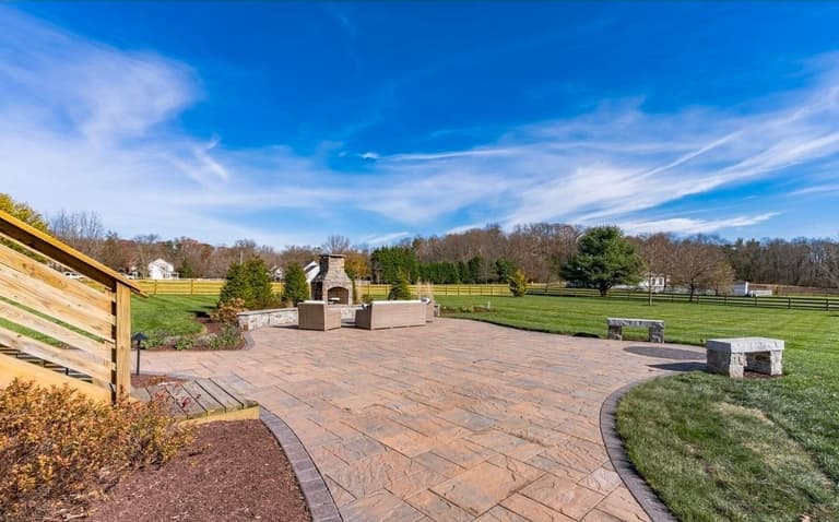 Large paver patio with a stone outdoor fireplace, seat wall and stone benches, seen from the deck stairs