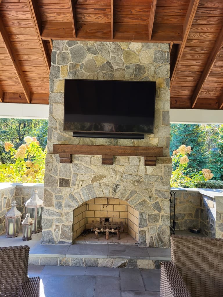 Stone outdoor fireplace with a mounted television and a reclaimed-timber mantel under a pavilion