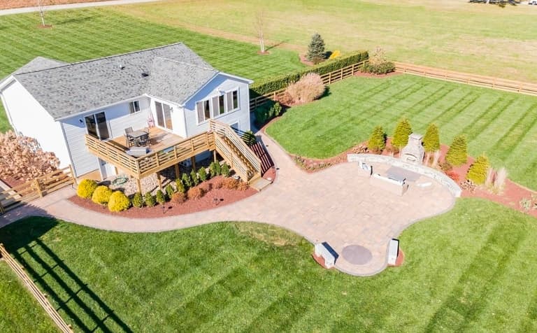 Aerial view of a curved paver patio linking a deck to a stone-fireplace terrace and a fire-pit seating area