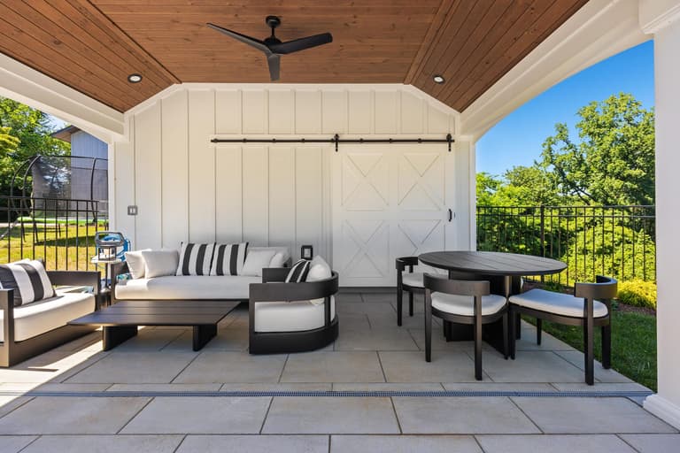 Covered pavilion with a cedar ceiling, white barn doors, and lounge and dining seating on a large-format paver floor