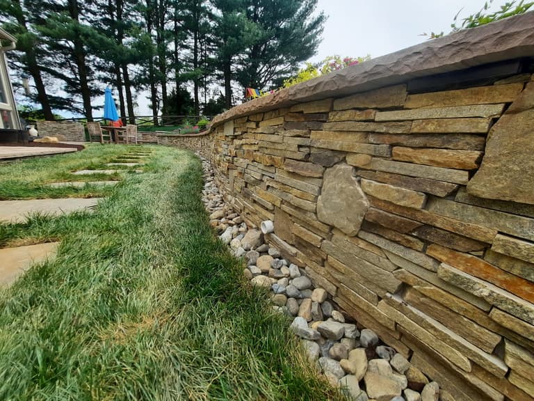 Curved stacked-stone retaining wall with a flagstone cap and a river-rock drainage bed at its base